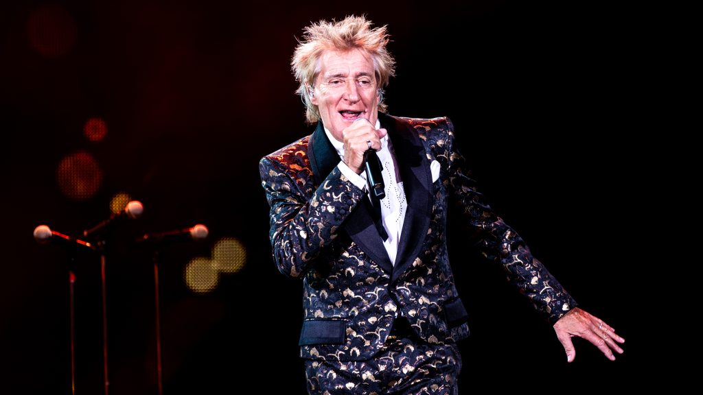 Rod Stewart sonriendo y señalando al público durante una actuación en vivo.
